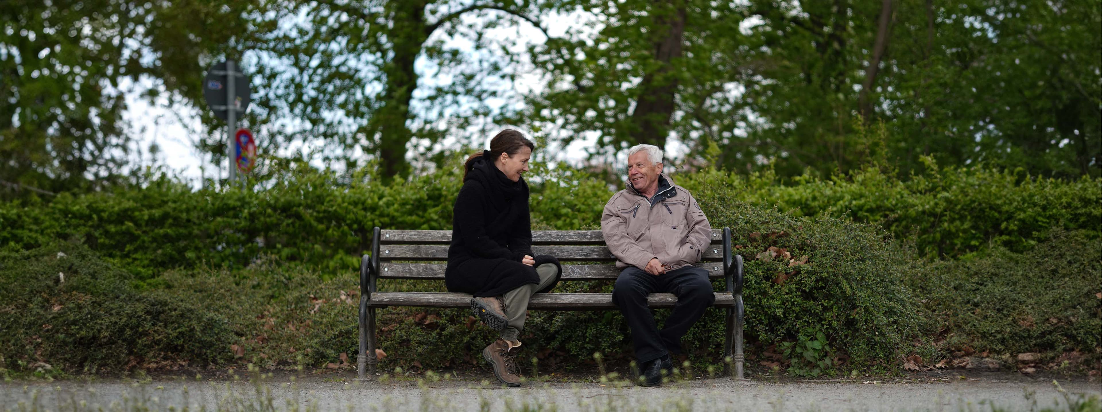 Silke Lezim und ein älterer Herr sitzten auf einer Parkbank und unterhalten sich